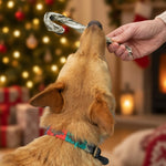A dog holding a Skipper's Fish Skin Candy Cane in its mouth while a human hand offers another ornament, with a blurred Christmas tree and cosy indoor background.